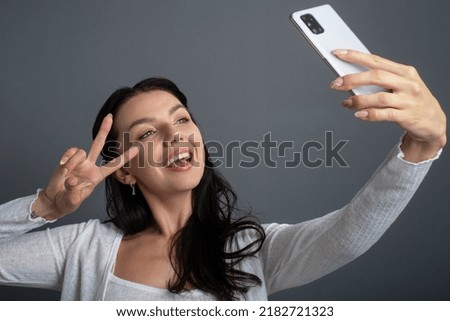 Taking selfie photos on cellphone. Young woman laughing and showing a victory peace gesture sign to a  front mobile phone camera.