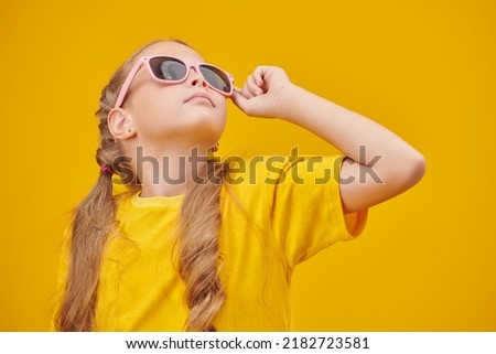 A stylish little blonde girl with pigtails poses in a bright yellow t-shirt and fashionable sunglasses looking up on a yellow background. Children's summer fashion. 