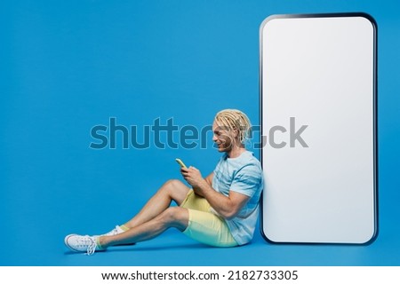 Full body young blond man with dreadlocks he in white t-shirt sit near big huge blank screen mobile cell phone workspace copy space area use smartphone isolated on plain pastel light blue background
