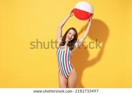 Happy young woman slim body wear striped red blue one-piece swimsuit hold play inflatable beach ball isolated on vivid yellow color wall background studio. Summer hotel pool sea rest sun tan concept