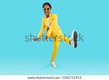 Full body shot of happy funny cheerful positive attractive young African American woman wearing stylish yellow suit and cool sunglasses dancing isolated on blue background. Party and fashion concept