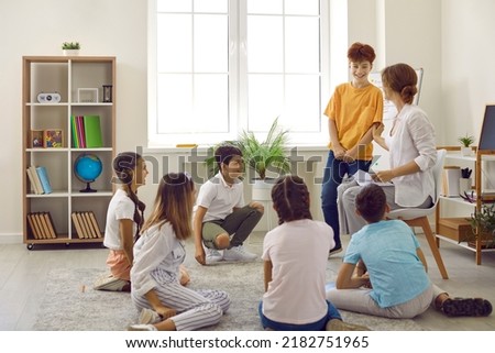 Children and teacher having circle time in class. Group of happy, cheerful school students sitting on the floor in a modern classroom interior, listening to interesting stories and discussing them