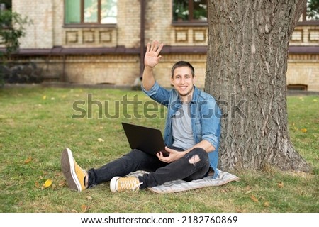 smiling emotional man outdoors looking aside waving hello. Young man with laptop rest in grass at university campus. Business man sit on lawn work outdoors on computer in a park. education concept