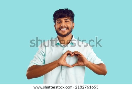 Portrait of joyful attractive indian guy showing love gesture on light blue background. Happy bearded young man in shirt with smile shows heart folded with fingers. Body language concept.