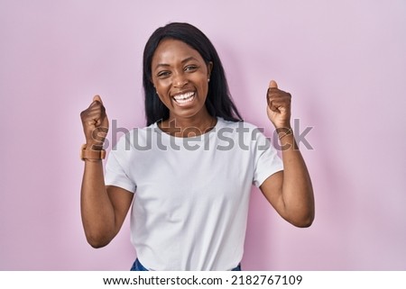 African young woman wearing casual white t shirt very happy and excited doing winner gesture with arms raised, smiling and screaming for success. celebration concept.  Royalty-Free Stock Photo #2182767109