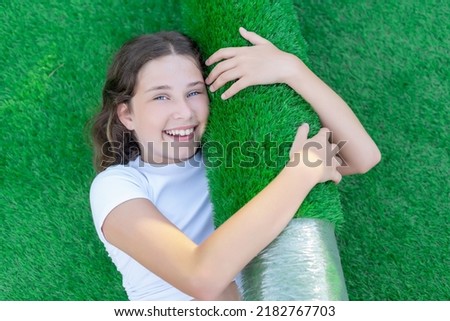 Smiling young caucasian girl lying on the lawn and hugging a new roll of artificial turf. Soft and squishy artificial grass ready to lay Royalty-Free Stock Photo #2182767703