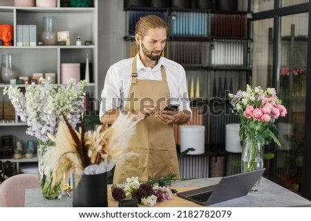 Small business. Young bearded male florist in flower shop writing a message to customer using smart phone. Floral design studio, making decorations and arrangements. Flowers delivery, creation order.