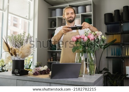 Small business. Attractive young male florist make bouquet and taking photo on smart phone for social media. Flowers delivery, order creation concept.