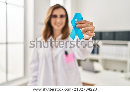 Young woman wearing doctor uniform holding blue ribbon at clinic