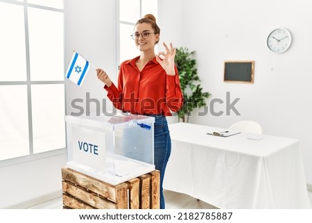 Young brunette woman voting putting envelop in ballot box holding israel flag doing ok sign with fingers, smiling friendly gesturing excellent symbol 