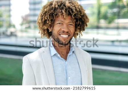 Close up photo of a handsome attractive positive african american or brazilian man with curly hair, dressed in stylish formal wear, standing outdoors, looking at the camera, smiling friendly
