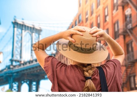 Fashionable tourist woman visiting New York City wearing hat and stylish street style outfit. Young girl amazed by the beautiful city. Manhattan bridge view on summertime.