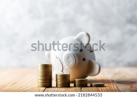 A sad piggy bank behind stacks of euro coins symbolizing the fall of monetary assets. White moneybox with coins graph. Crisis concept Royalty-Free Stock Photo #2182844533