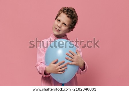 beautiful cute little boy of school age stands with a blue balloon in his hand and poses smiling broadly standing in a pink shirt Photo on an empty pink background with space for an advertising insert