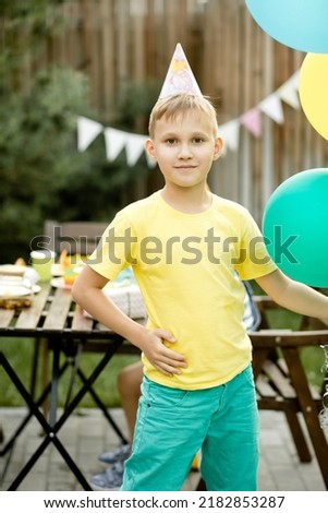 Cute funny nine year old boy celebrating his birthday with family or friends in a backyard. Birthday party. Kid wearing party hat and holding balloon in a hand.