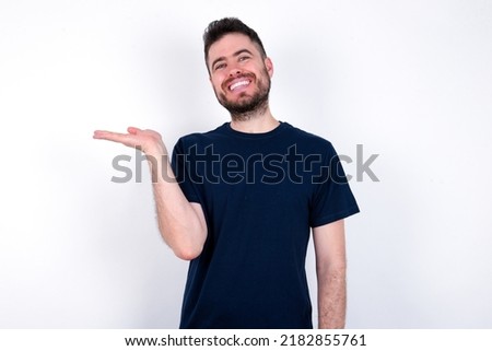Young caucasian man wearing black T-shirt over white background smiling cheerful presenting and pointing with palm of hand looking at the camera.