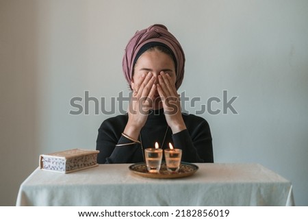 Jewish woman prays over lit Shabbat candles, covering her face with her hands. Nearby lies a religious prayer book. Jewish religious traditions Royalty-Free Stock Photo #2182856019