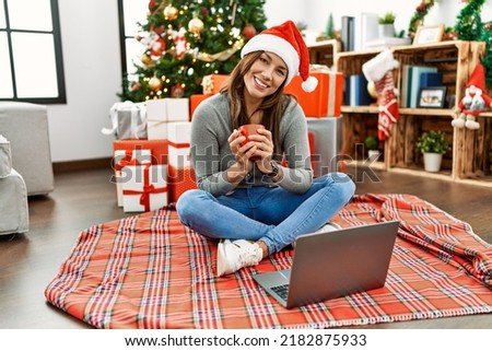 Young latin woman watching movie and drinking coffee sitting by christmas tree at home