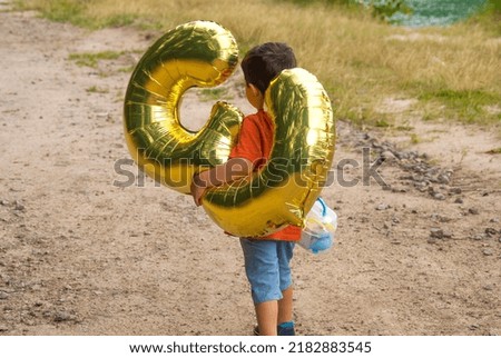 A child walks down the road carrying an inflatable golden balloon in the form of the number three in one hand and a suitcase with toys in the other hand