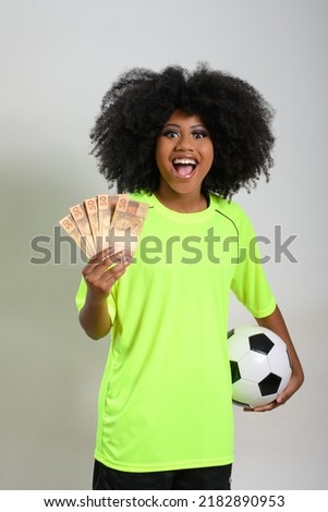 woman holding brazilian money is a soccer ball, she wears neon green uniform, isolated on gray background
