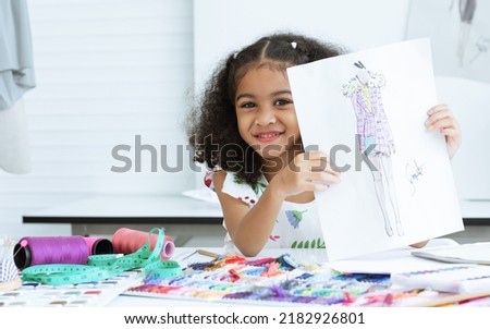 Cute African little girl showing, drawing, sketching clothing design on paper at tailor room, cozy home as her career dream, smiling with happiness and fun. Leisure, Lifestyle, Education, Kid Concept