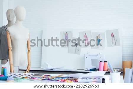 Background of interior tailor shop or room with nobody, there are mannequin, clothes, thread, sewing machine on table and paper of clothing design on wall. Lifestyle and small business concept Royalty-Free Stock Photo #2182926963