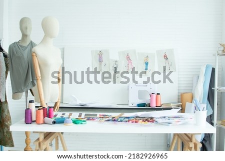 Background of interior tailor shop or room with nobody, there are mannequin, clothes, thread, sewing machine on table and paper of clothing design on wall. Lifestyle and small business concept Royalty-Free Stock Photo #2182926965