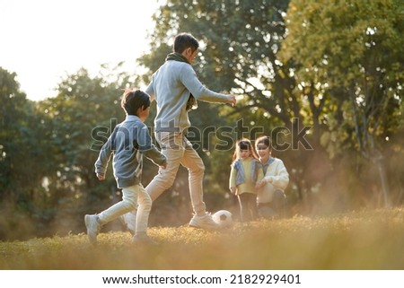 asian father and son playing soccer football outdoors in park while mother and daughter watching on the side