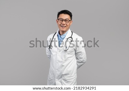 Cheerful confident handsome middle aged asian male therapist in white coat, glasses with stethoscope look at camera isolated on gray background. Disease treatment, health care, medicine, offer and ad Royalty-Free Stock Photo #2182934291