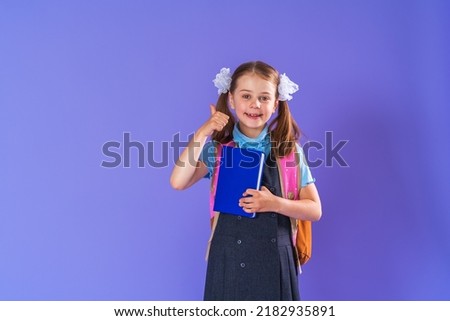 happy schoolgirl in uniform with a backpack, standing on a purple background in the studio with book in her hands. the little girl is ready for school and shows thumbs up gesture, sign of approval.