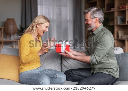 Loving middle aged husband surprising his wife with gift at home, greeting her with birthday or anniversary, happy man giving present box to excited spouse, celebrating valentine's day together