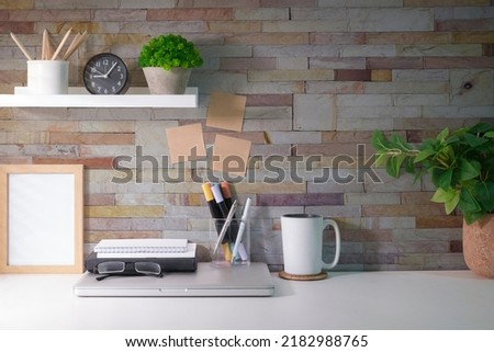 Home office desk with supplies, picture frame, house plant and coffee cup on white table.