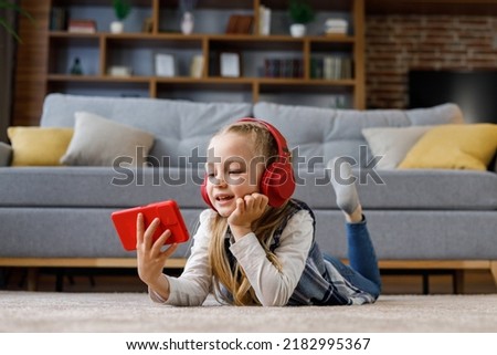 Little girl wearing red earphones lying on the carpet at home. Cute smiling child holding smartphone, watching cartoons, browsing internet or watching video in social media, playing online games