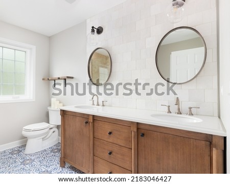 A cozy bathroom with a patterned tile floor, natural wood vanity, tiled backsplash, and lights mounted above circular mirrors. Royalty-Free Stock Photo #2183046427