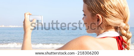 Panoramic rear view of a young attractive woman on holiday laying down on a beach by the sea, holding and using a smartphone to take pictures of the scenery. People travel technology outdoors.