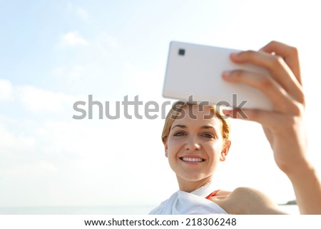 Close up of attractive young tourist woman relaxing on a sandy beach by the sea, holding a smartphone device taking selfies pictures of herself, networking on holiday. People technology outdoors.