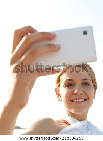 Close up of attractive young tourist woman relaxing on a sandy beach by the sea, holding a smartphone device taking selfies pictures of herself, networking on holiday. People technology outdoors.