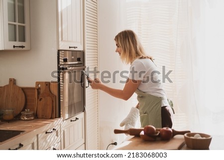 charming young woman in chef's apron puts apple pie in the oven and takes pictures on phone
