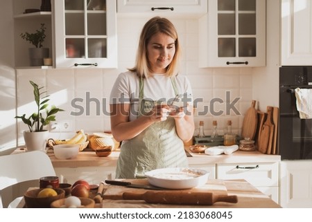 Pretty young woman preparing apple pie at home in cozy kitchen and takes pictures on phone
