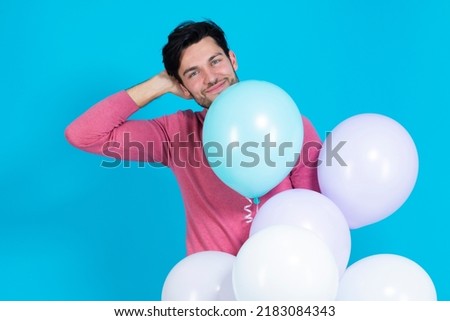 One Caucasian Guy Handsome Brunet Man With Bunch of Colorful Air Balloons in Pink Jumper Standing With Lifted Hand On Blue Background. Horizontal image Composition