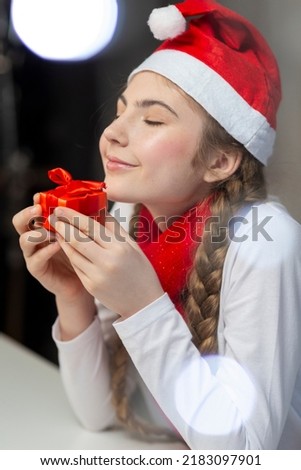 One Dreaming Cute Winsome Caucasian Female Girl in Santa Hat and White Shirt With Tiny Red Giftbox Relaxing and Thinking. Vertical Image