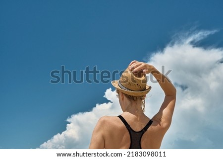 Woman holds a straw hat on her head with her hand, a silhouette of a beautiful blonde against the background of cumulus clouds over the sea Royalty-Free Stock Photo #2183098011