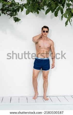 full length of athletic man in swimming trunks and sunglasses posing near white wall Royalty-Free Stock Photo #2183100187