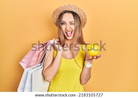 Young blonde girl holding shopping bags and piggy bank sticking tongue out happy with funny expression. 
