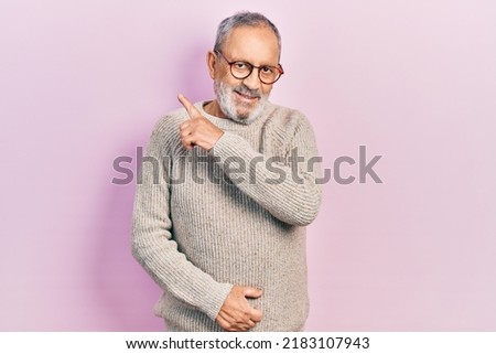 Handsome senior man with beard wearing casual sweater and glasses cheerful with a smile on face pointing with hand and finger up to the side with happy and natural expression 