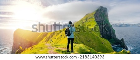 Happy woman hiker using smartphone taking photo at coast cliff edge during travel Faroe Island. Tourist admire of Kallur Lighthouse, Kalsoy island. Panoramic photo during sunset or sunrise.  Royalty-Free Stock Photo #2183140393