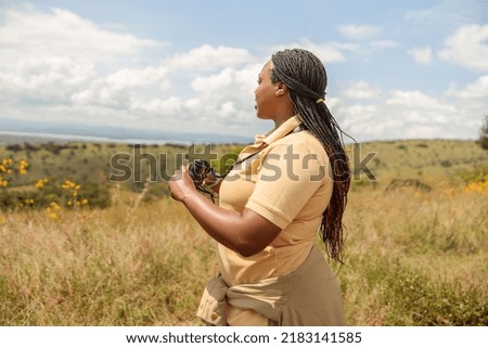 Side view of African American professional female guide searching the landscape for wildlife in the savannah, holding binoculars Royalty-Free Stock Photo #2183141585
