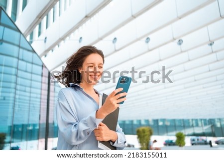 Confidence businesswoman female with smartphone texting message while standing outdoors in the city business district. Young smiling woman using mobile app near modern office building. Technology Royalty-Free Stock Photo #2183155011