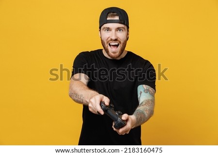 Young cheerful bearded tattooed man 20s he wears casual black t-shirt cap hold in hand play pc game with joystick console isolated on plain yellow wall background studio portrait. Tattoo translate fun