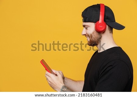 Close up side view fun young happy bearded tattooed man 20s he wearing casual black t-shirt cap headphones listen to music dance use mobile cell phone isolated on plain yellow wall background studio. Royalty-Free Stock Photo #2183160481